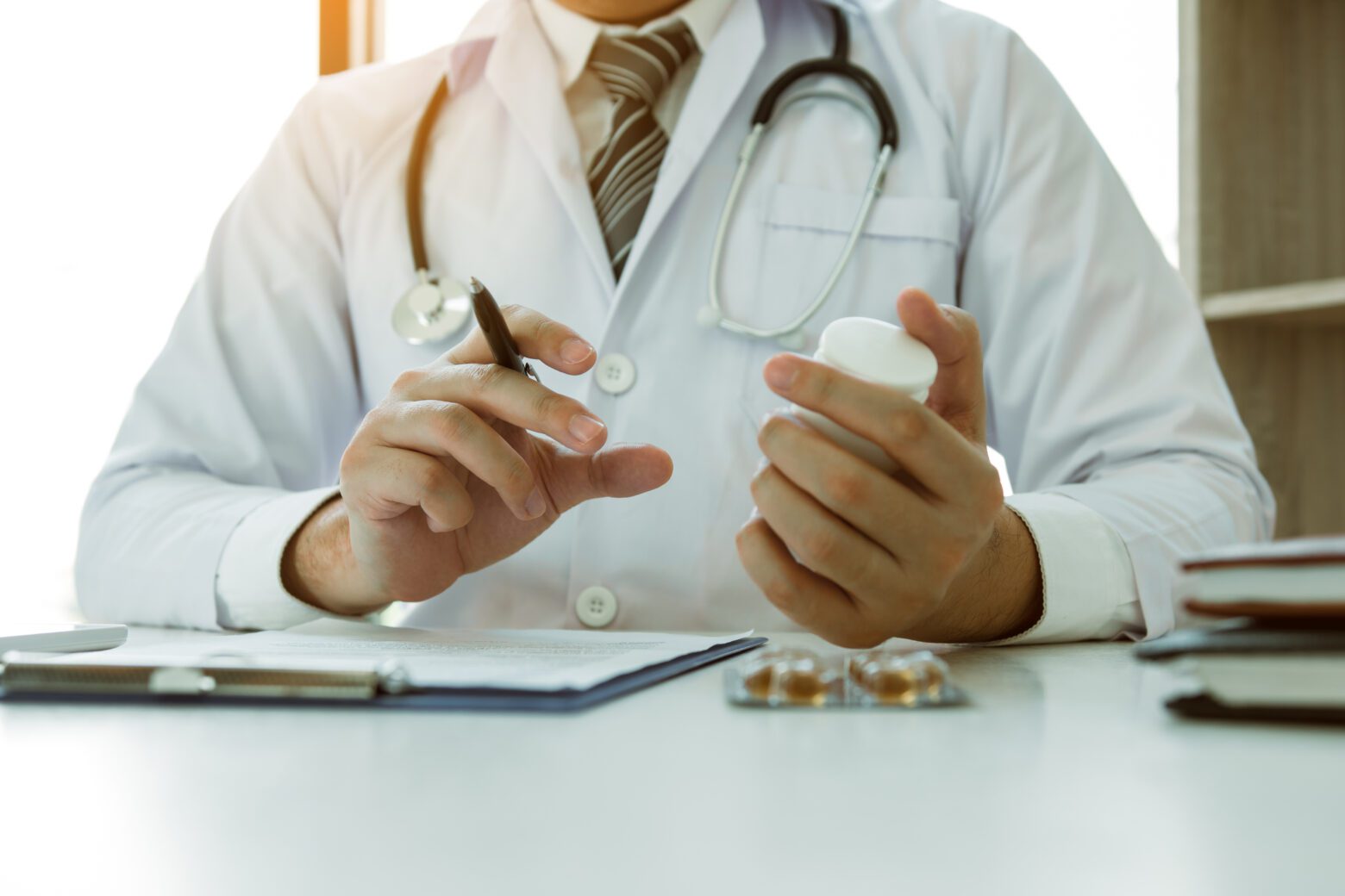 Confident doctor man holding a pill bottle and writing while talking with senior patient and reviewing his medication at office room.